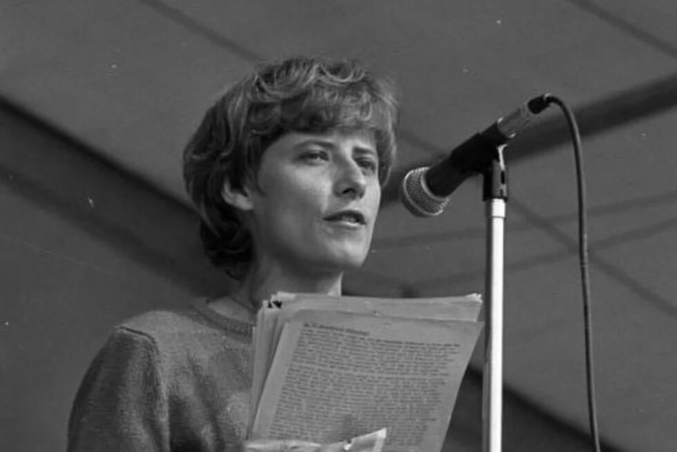 Schwarz-weiß Foto von Petra Kelly, die mit einem Manuskript in den Händen am Mikrofon steht