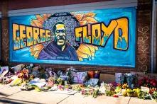 Grafitti auf dem George Floyd abgebildet und geschrieben ist, im Vordergrund liegen viele Blumen  und Plakate