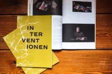 Drei Ausgaben des Magazins Interventionen liegen auf einem Holzdielenboden, eines der Magazine ist aufgeschlagen.