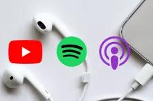 Ein Smartphone mit weißen Kopfhörern liegt auf einem Tisch. Darauf sind die Symbole für Youtube, Spotify und Apple Podcasts.