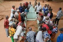 Screenshot aus dem Film "Das leere Grab": Man sieht Schwarze Menschen, die um einen Sarg herumstehen, der komplett mit Sand gefüllt ist.