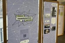 Foto von den Ausstellungs-Bannern: Sie haben einen blass-lila Hintergrund mit Text und Foots