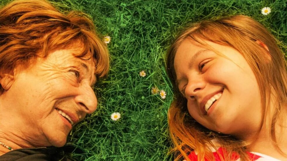 Foto aus dem Film "Wiedersehen mit Brundibar" von Douglas Wolfsperger. Eine ältere Frau und ein Mädchen liegen auf einer Wiese, ihre Köpfe liegen dicht beieinander und sie sehen sich lachend an.