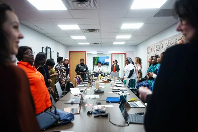Photo: People* stand in a conference room around a long table with laptops and papers. In the background, two people in front of a whiteboard.
