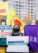 Drei Menschen auf einer Demonstration, sie haben Masken auf und Plakate in der Hand.